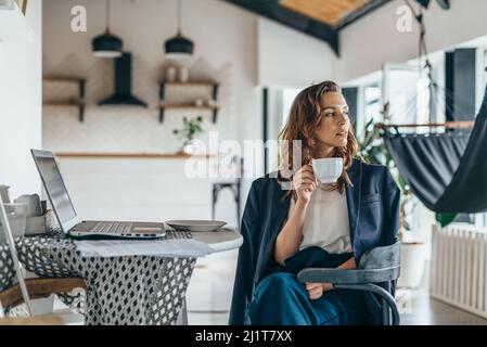 A woman is sitting in the kitchen, distracted from working with her laptop, drinking from a mug and looking thoughtfully away. Stock Photo