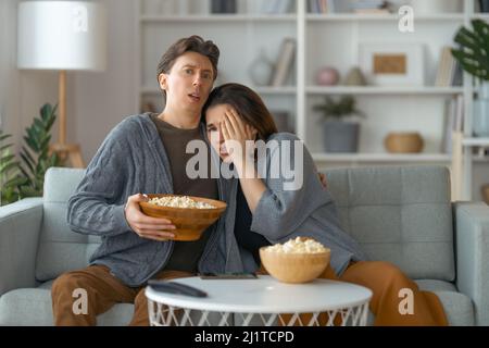 Shocked family with popcorn sitting on sofa and watching TV at home. Couple spending time together. Stock Photo