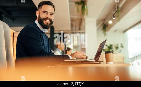 businessman working on his laptop in a co-working space. Modern businessman smiling at the camera while typing on his laptop and listening to music. H Stock Photo