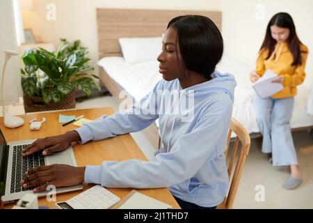 Pensive Black college student working on laptop when her friend reading notes in notebook in background Stock Photo