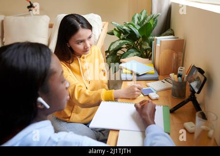 Serious high school students sitting on floor in front of smartphone when attending online class Stock Photo