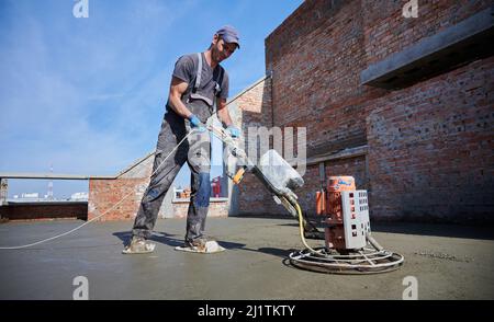 Young builder in workwear working with grout-grinding machine for alignment and smoothing floor surface. Worker grouting floor before applying finish coverings. Stock Photo