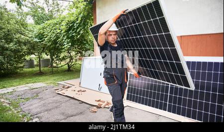 Serious man solar installer carrying solar module in the yard. Male worker in work overalls holding photovoltaic solar panel and walking down asphalt pathway outside the house. Stock Photo