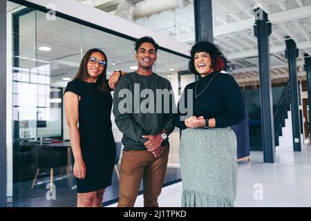 Happy colleagues smiling at the camera while standing together in a modern office. Team of three businesspeople wearing business casual in a creative Stock Photo
