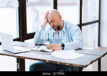 Portrait of tired middle-aged bald man wearing blue shirt, sitting at wooden desk near documents, laptop in office. Stock Photo