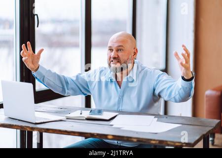 Portrait of angry annoyed middle-aged bald man wearing blue shirt sitting at wooden desk near document laptop in office. Stock Photo