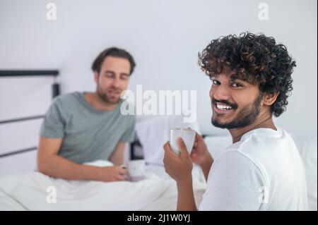Man drinking coffee with his sleepy boyfriend in the bedroom Stock Photo