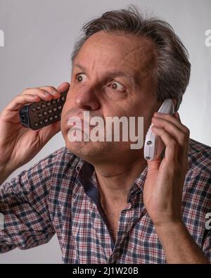 A man with a shocked expression listens to two phone calls from two different cordless phones which he holds one in each ear Stock Photo