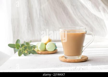 a transparent cup of coffee with milk with macaroons and eucalyptus leaves at morning shadows and sun rays through the window on gray table background Stock Photo
