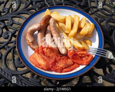 Lunch outdoors with traditional pork sausages, grilled plum tomatoes and potato chips on  blue edged white plate Stock Photo