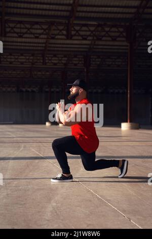 Full body side view of muscular male in sporstwear and cap doing lunges on sunny street during training in city Stock Photo