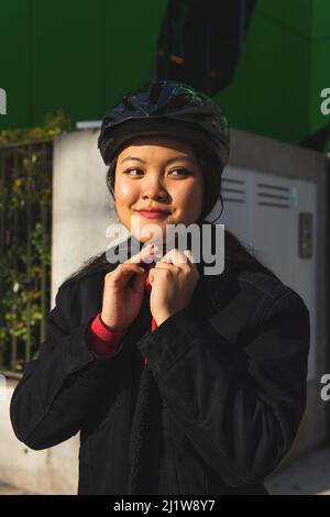 Charming young Asian female in casual outfit fastening helmet while preparing for practicing activity on spare time in park Stock Photo