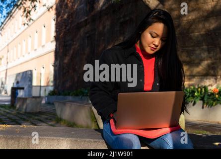 Serious ethnic female teleworker in casual clothes sitting on stone bench and typing on netbook while working remotely Stock Photo