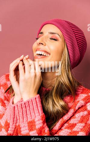 Beautiful young Woman with makeup in warm sweater standing on pink background with closed eyes while touching forehead in studio Stock Photo