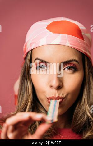 Attractive female with makeup eating sour colorful candy strip and looking at camera while standing on pink background in light studio Stock Photo
