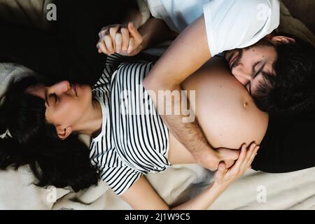 Top view of gentle loving husband hugging belly of pregnant wife while relaxing on soft bed at home Stock Photo