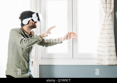 Young bearded male with dark hair in casual clothes gesturing and smiling while experiencing virtual reality in modern headset standing near window at Stock Photo