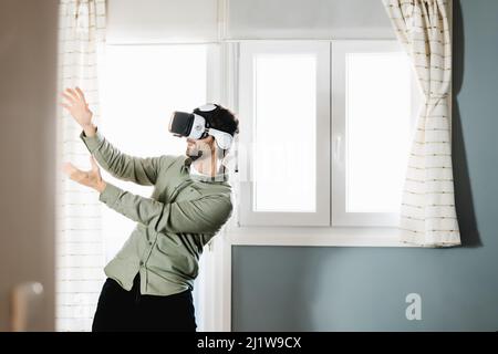 Young bearded male with dark hair in casual clothes gesturing and smiling while experiencing virtual reality in modern headset standing near window at Stock Photo