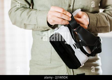 Cropped unrecognizable attentive young ethnic man with dark hair and beard in casual clothes examining VR goggles while standing at home in daylight Stock Photo