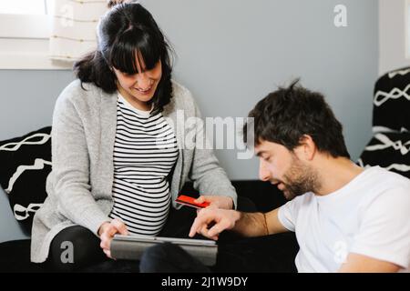 Cheerful young pregnant Hispanic Woman with dark hair in casual clothes smiling and looking at husband while using tablet together on comfortable sofa Stock Photo