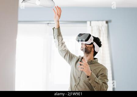 Young bearded male with dark hair in casual clothes gesturing and smiling while experiencing virtual reality in modern headset standing near window at Stock Photo