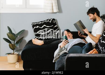 Young calm ethnic pregnant Woman in casual clothes messaging on smartphone while lying on knees of bearded husband using tablet on sofa Stock Photo