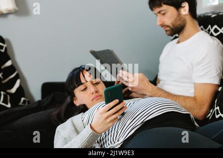 Young calm ethnic pregnant Woman in casual clothes messaging on smartphone while lying on knees of bearded husband using tablet on sofa Stock Photo