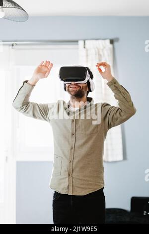 Young bearded male with dark hair in casual clothes gesturing and smiling while experiencing virtual reality in modern headset standing near window at Stock Photo