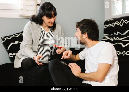 Cheerful young pregnant Hispanic Woman with dark hair in casual clothes smiling and looking at husband while using tablet together on comfortable sofa Stock Photo