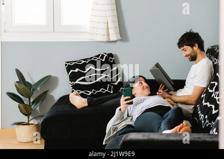 Young happy calm ethnic pregnant Woman in casual clothes messaging on smartphone while lying on knees of bearded husband using tablet on sofa Stock Photo