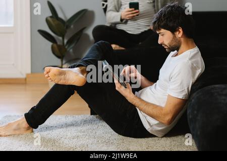 Bearded barefoot man leaning on sofa and scrolling tablet near cropped unrecognizable pregnant Woman using cellphone in living room Stock Photo