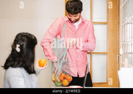 Side view of young anonymous female freelancer sitting at table and working on laptop while husband standing in kitchen with net sack with assorted fr Stock Photo