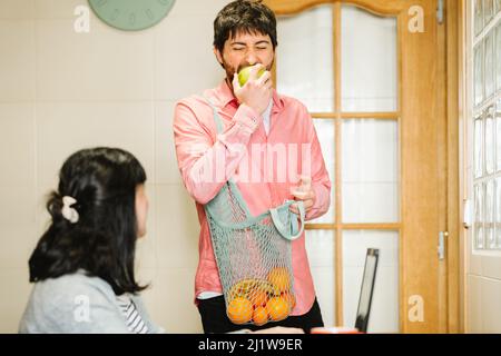 Young Hispanic man with dark hair eating fresh green apple to wife sitting at table and working remotely on laptop Stock Photo