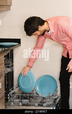 Young ethnic bearded male with dark hair in shirt putting plates into dish washing machine in kitchen Stock Photo