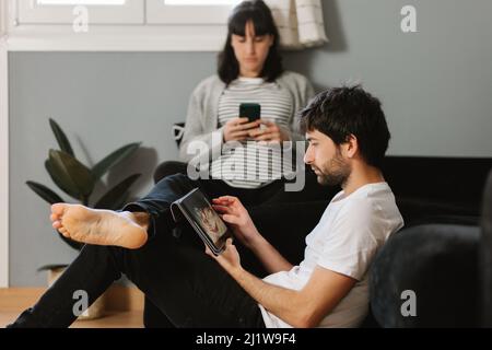 Bearded barefoot man leaning on sofa and scrolling tablet with pregnant Woman using cellphone in living room Stock Photo