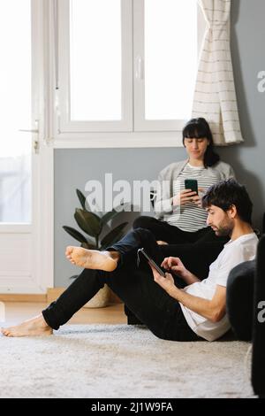 Bearded barefoot man leaning on sofa and scrolling tablet with pregnant Woman using cellphone in living room Stock Photo