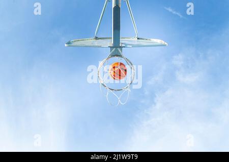 From below of orange basketball falling into round hoop with net placed against blue sky on sunny summer day in city Stock Photo