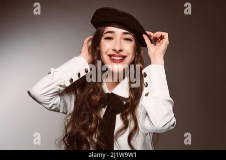 Glad Hispanic Woman with long wavy hair and makeup wearing trendy headgear and looking at camera while putting on his beret against degraded backgroun Stock Photo