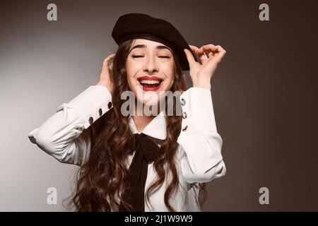 Glad Hispanic Woman with long wavy hair and makeup wearing trendy headgear and looking at camera while putting on his beret against degraded backgroun Stock Photo