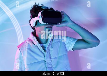 man in modern black VR goggles experiencing virtual reality while standing near white wall with glowing lights in contemporary studio Stock Photo