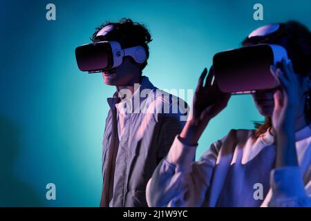 Couple in casual wear and modern VR goggles exploring virtual reality together while standing near wall in studio with dim illumination Stock Photo