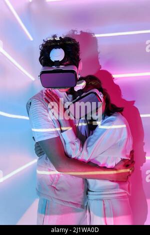 Front view of couple embraces in modern VR goggles exploring cyberspace at wall with bright glowing lights in studio Stock Photo