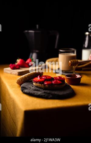 Crop anonymous person with sweet honey on wooden spoon for bagel with slices of fresh strawberries on chocolate paste on table with coffee from moka p Stock Photo