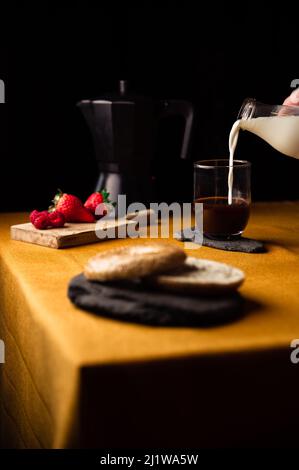 Crop anonymous person pouring milk from jug into glass cup with coffee brewed in moka pot for breakfast with strawberries and bagel on slate plate Stock Photo