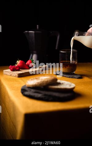 Crop anonymous person pouring milk from jug into glass cup with coffee brewed in moka pot for breakfast with strawberries and bagel on slate plate Stock Photo