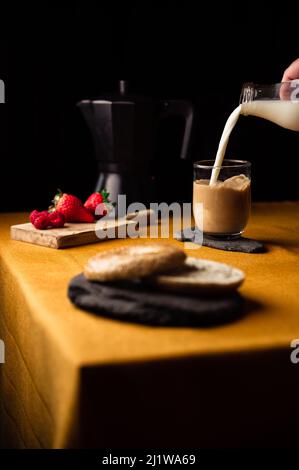 Crop anonymous person pouring milk from jug into glass cup with coffee brewed in moka pot for breakfast with strawberries and bagel on slate plate Stock Photo