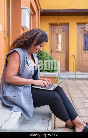 Side view of concentrated African American female freelancer in formal wear sitting on stairs and typing on netbook while working remotely Stock Photo