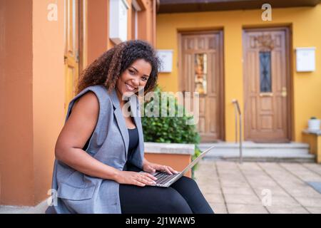 Happy African American female freelancer in formal wear sitting on stairs and typing on netbook while working remotely Stock Photo