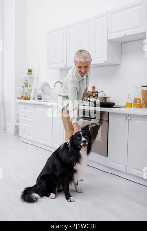 Positive senior woman petting border collie while cooking in kitchen Stock Photo