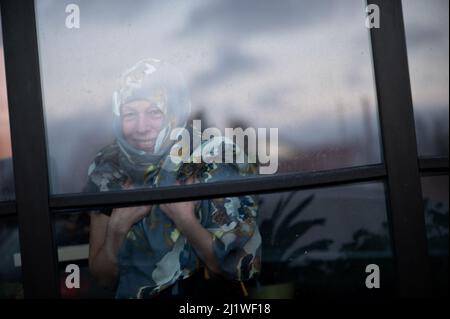 Smiling middle-aged woman with scarf on head looking through dirty window glass Stock Photo